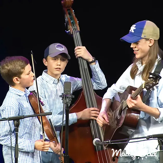Children performing in an indoor auditorium with musical instruments, stage lighting, and dark background.
