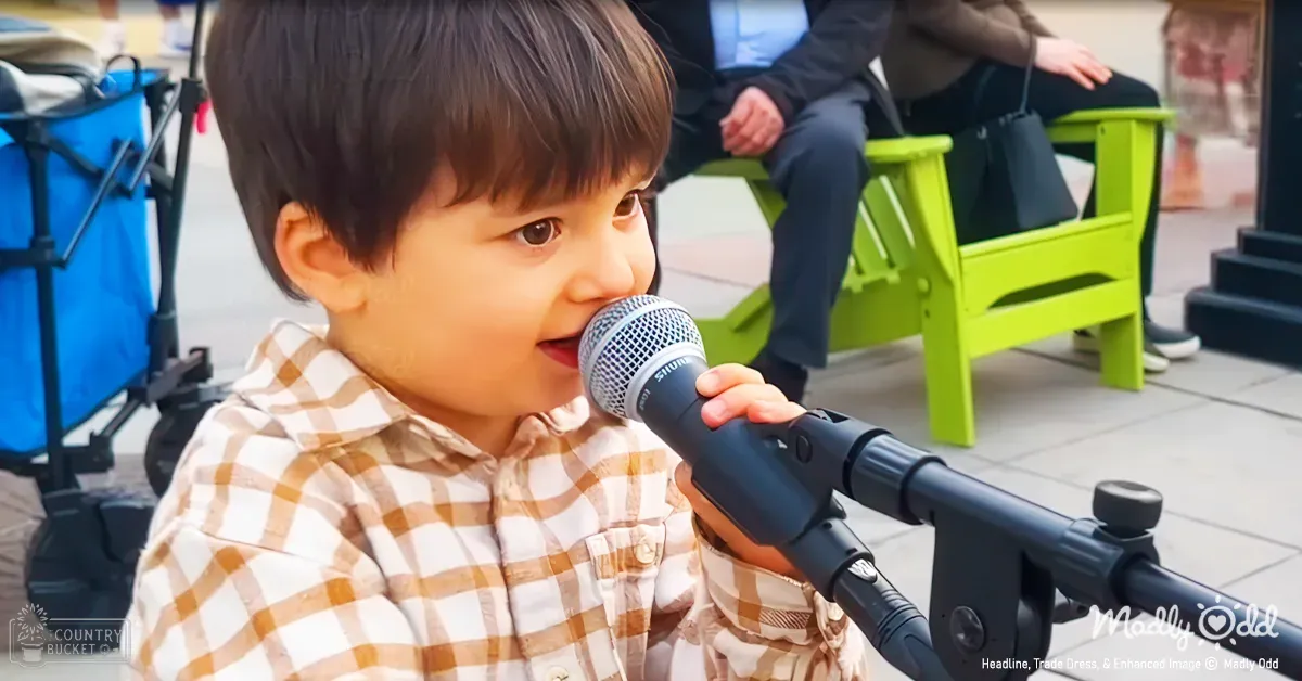 A child sings into a microphone outdoors with green chairs and blue bag in the background, suggesting spring or summer.