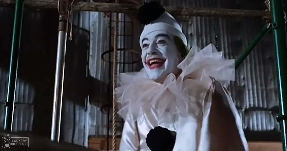 An adult male clown wearing a white ruffled collar shirt and black hat with a pom-pom, holding onto a rope or cord in a dimly lit industrial backstage area of a theater.