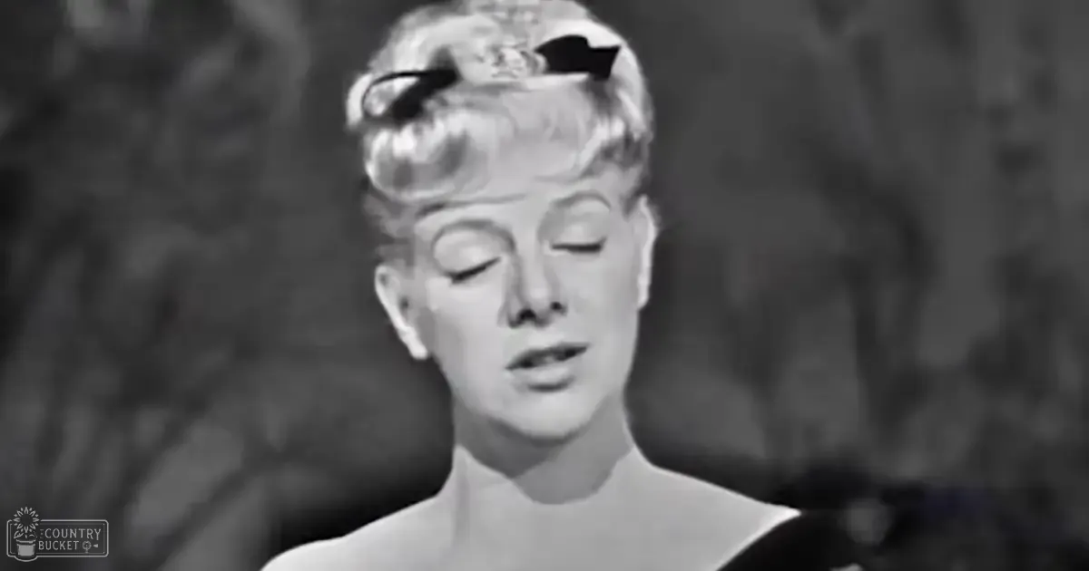 A woman wearing a sleeveless top with a high neckline stands on an ornate stage backdrop, possibly from the mid-20th century theater setting.