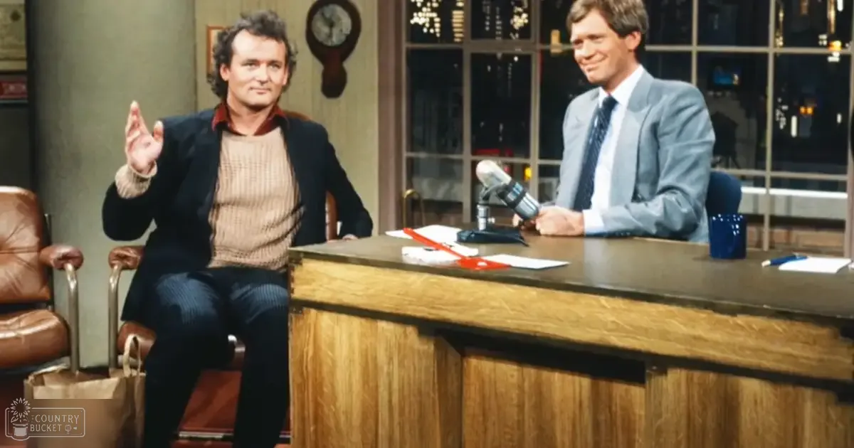 An adult male singer wearing a dark blazer over a beige sweater vest and dark patterned trousers, seated on a late-night talk show set with another person in a light gray suit.