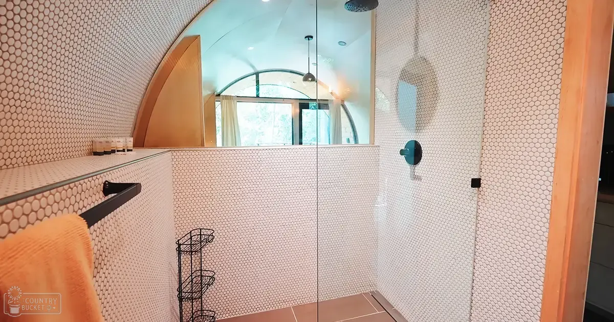 A modern bathroom interior with a glass-enclosed shower, hexagonal tiles in beige and white, an orange towel hanging nearby, and black metal fixtures.