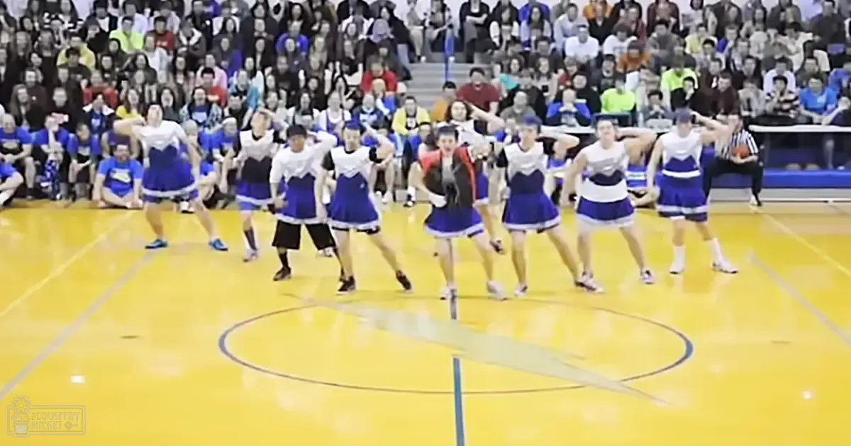 A cheerleading team performs at an indoor sports event, with a referee and seated audience visible. The setting is an arena or gymnasium with yellow court flooring.