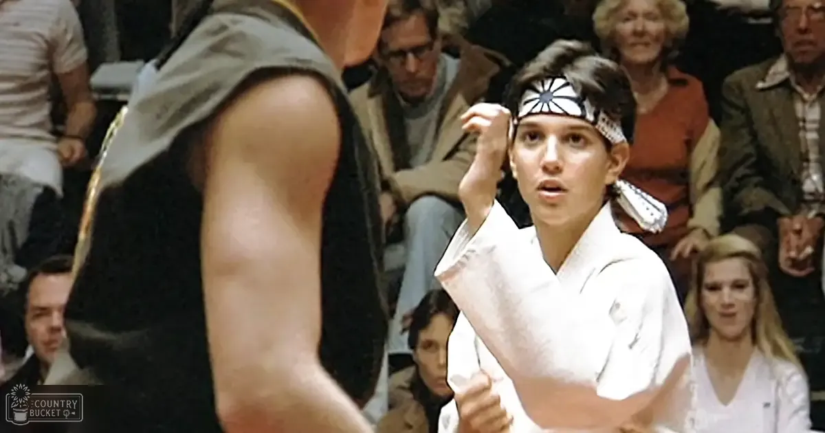 A young adult male wearing a white karate gi with black trim and headband, standing indoors, possibly in a gymnasium or dojo, watched by an audience of adults in various clothing suggesting cooler weather.