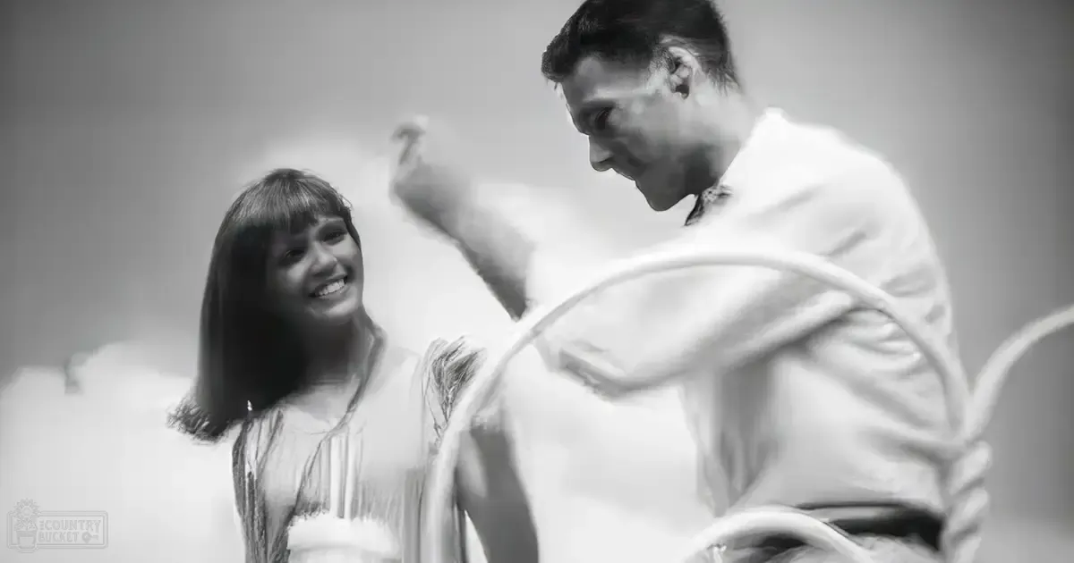 A young adult female in a light-colored striped dress stands beside a middle-aged male wearing a long-sleeved robe in an indoor setting with soft, ethereal lighting.