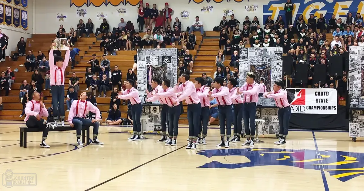 A dance group of teenagers in pink shirts and red suspenders performing at a school event in a gymnasium with bleachers filled with spectators.