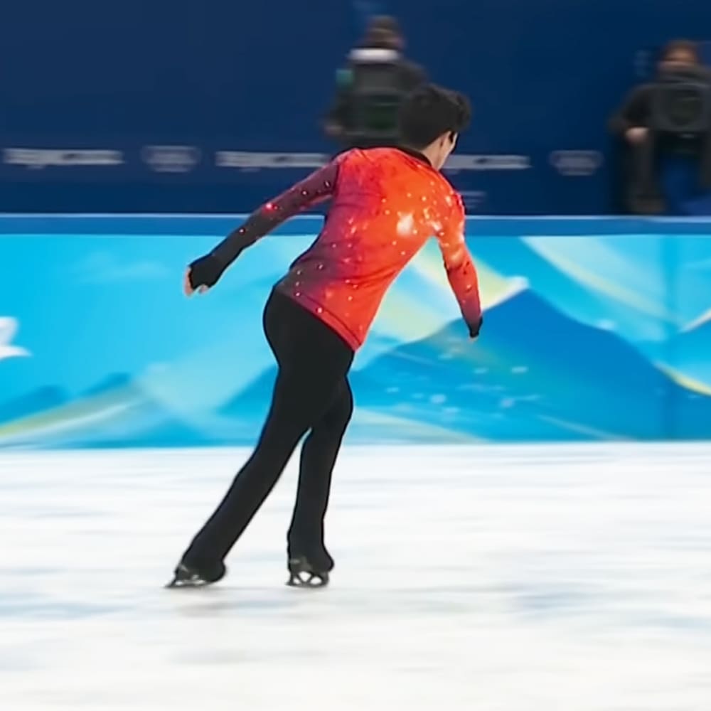 A young adult male figure skater in a red, star-patterned skating costume stands on an indoor ice rink with a blue backdrop with abstract designs. The scene is set under artificial lighting typical of an indoor sports venue.