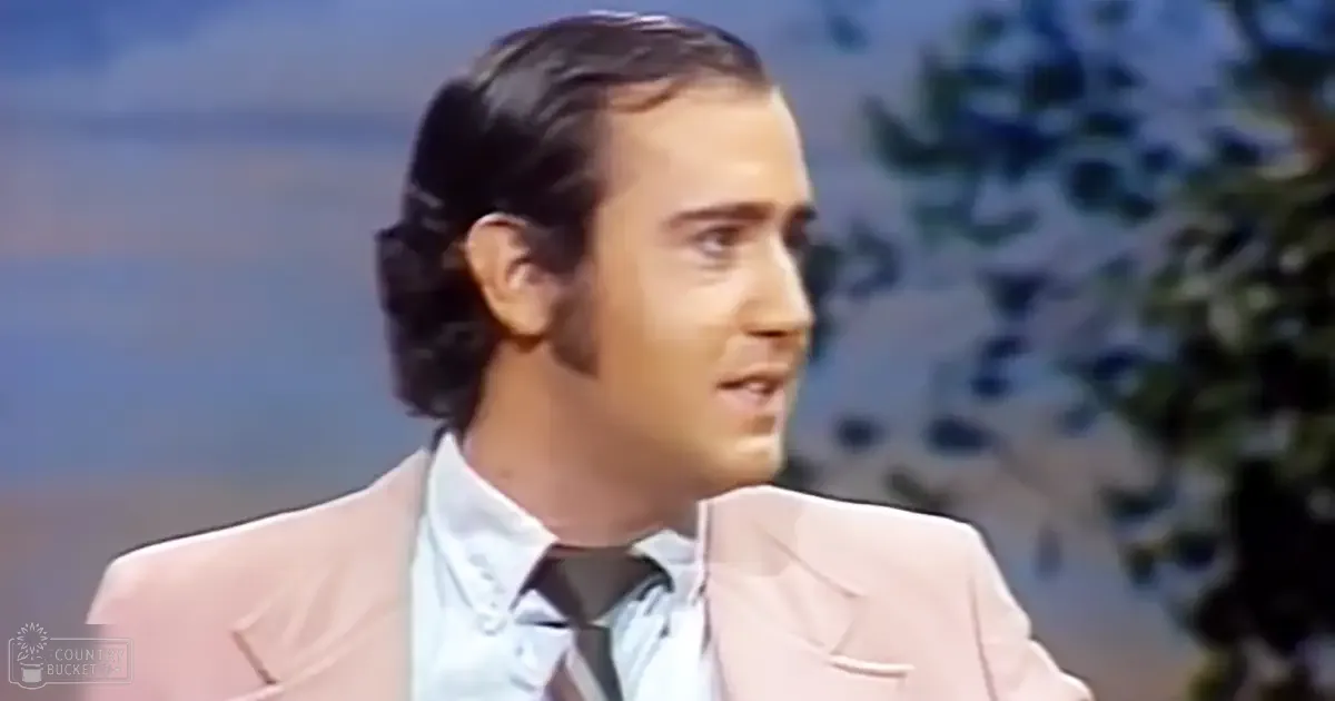An adult male in a tailored light pink suit jacket and white dress shirt, against an outdoor backdrop with foliage and soft afternoon light.