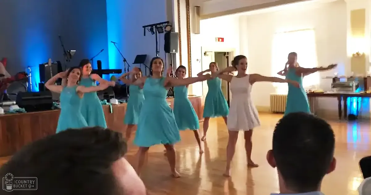 A dance performance with performers in turquoise dresses, an audience of two adults, and a setting suggesting daytime at an indoor venue.