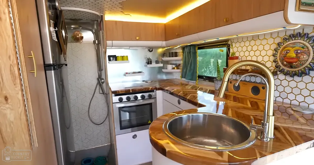 Compact kitchen setup in an RV or camper van, with stainless steel sink, wooden countertops, small oven, decorative wall tiles, and natural light during daytime.