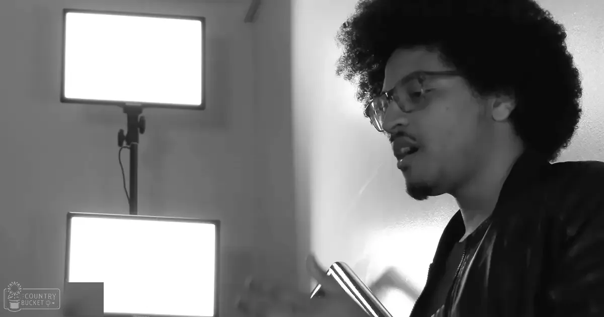 A young adult male wearing a leather jacket with glasses in an indoor setting, possibly a recording studio with bright light panels and minimal decor.