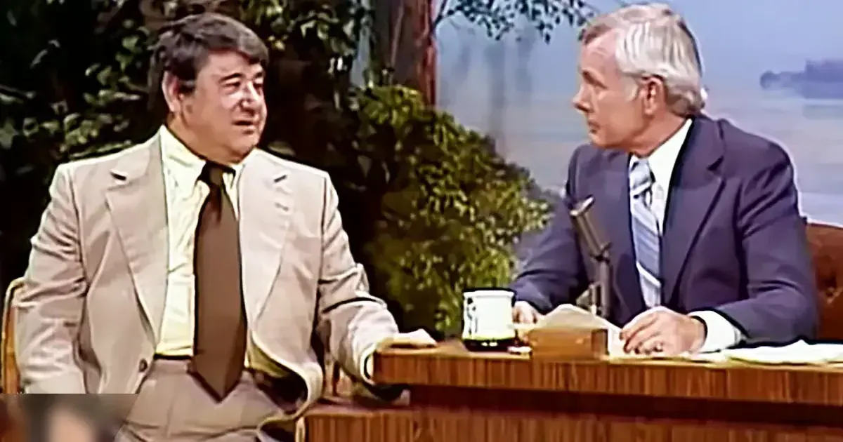 A talk show host with an older adult male guest in front of him; both wearing formal suits typical of the late 20th century, seated at a wooden desk with a microphone and papers, against a backdrop of blurred greenery and water suggesting a studio setting.