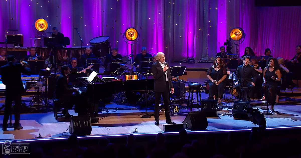An adult male singer in a dark suit performing at an indoor concert hall with bright stage lights and an orchestra seated behind him playing various instruments.