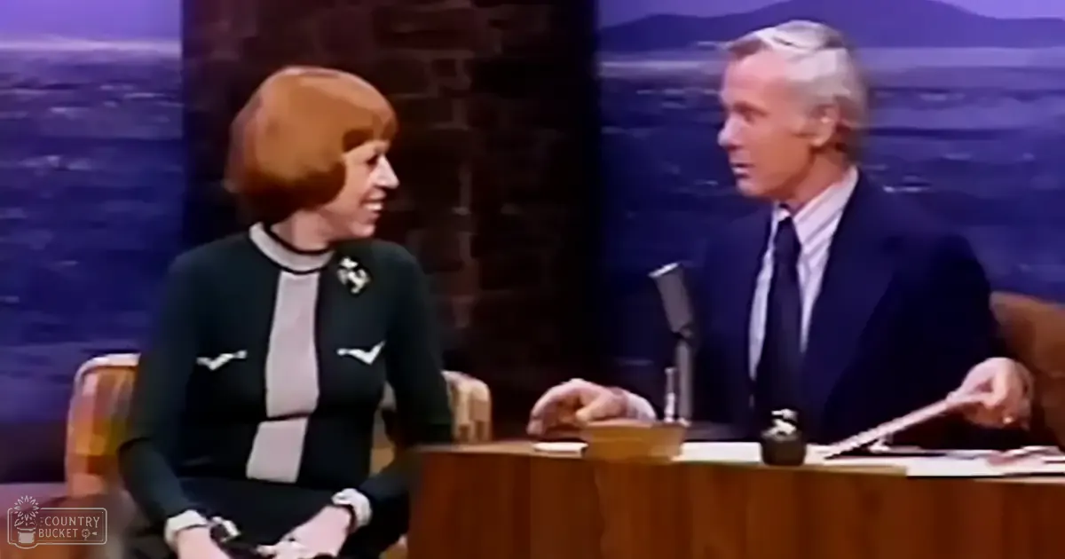 A late-night talk show setting with two hosts against a nighttime cityscape backdrop, with a female guest in a dark green blazer and male host in a classic dark suit.