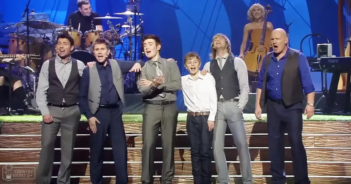 A musical performance group on stage with professional lighting and blue backdrop; various performers wearing button-down shirts and vests, including a child in white shirt.