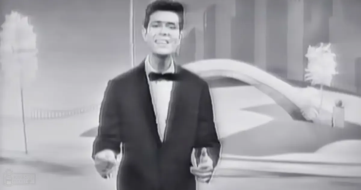 A young adult male wearing a formal mid-20th-century tuxedo with bow tie, indoors with a car adorned with white flowers in the background.