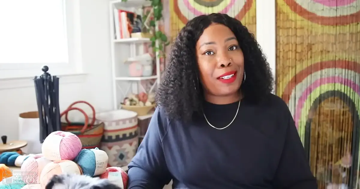 A woman in an indoor setting wearing a dark blue long-sleeved top with a silver necklace, surrounded by books, decorative items, yarns, and a colorful woven wall hanging.