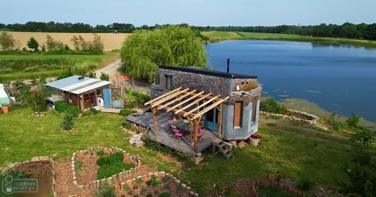 A rural landscape with a modern eco-friendly house overlooking a serene lake surrounded by green fields and trees on a clear daytime in spring or summer.
