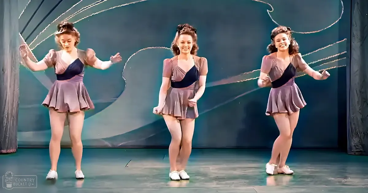 A dance performance scene with young adult female dancers in matching dark blue and light purple satin or silk outfits on a stage with abstract lighting effects.