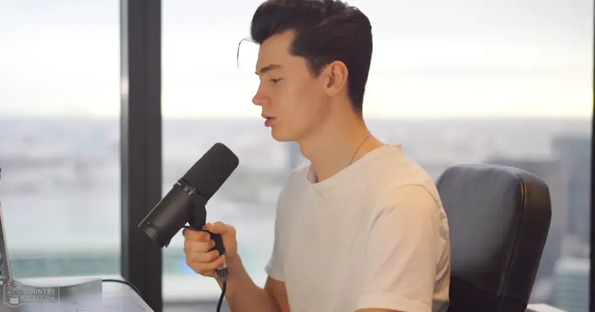 A young adult male wearing a white t-shirt and holding a black microphone sits at an office desk with a laptop, in front of large windows showing an urban cityscape. The setting appears to be during late afternoon or early evening.