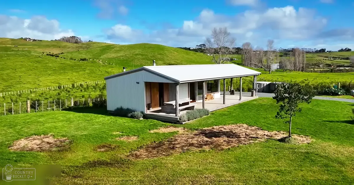 A small white and gray house with a porch, surrounded by green grass and hills under a clear blue sky in a rural setting.