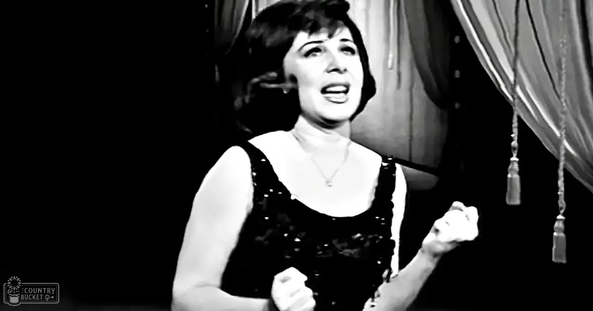 A woman wearing a black dress with a sweetheart neckline stands indoors under stage lighting.