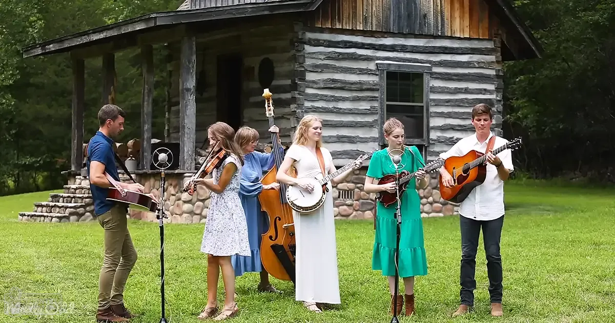 A small group performing music in front of a log cabin amidst greenery, suggesting an outdoor rural setting with natural light.