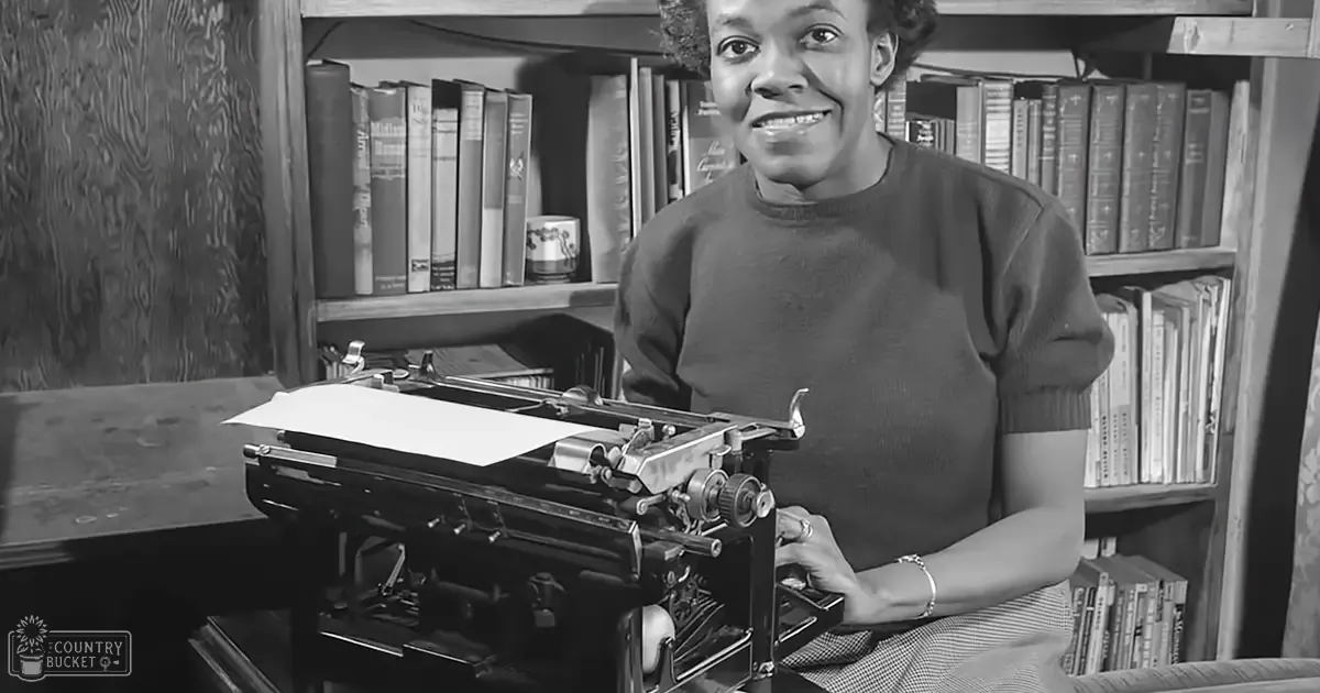 An adult female wearing a dark-colored, short-sleeved crew-neck sweater in an indoor setting with a wooden desk, bookshelf filled with books, and a typewriter.