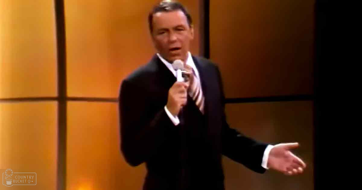 A middle-aged adult male performer in a classic fit dark suit with white shirt, holding a microphone, set against a warm-toned studio backdrop.