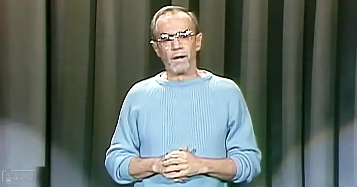 Older adult male in a studio setting, wearing a relaxed light blue long-sleeved ribbed sweater with thick-framed eyeglasses, simple vertical blinds indicating an indoor daytime environment.