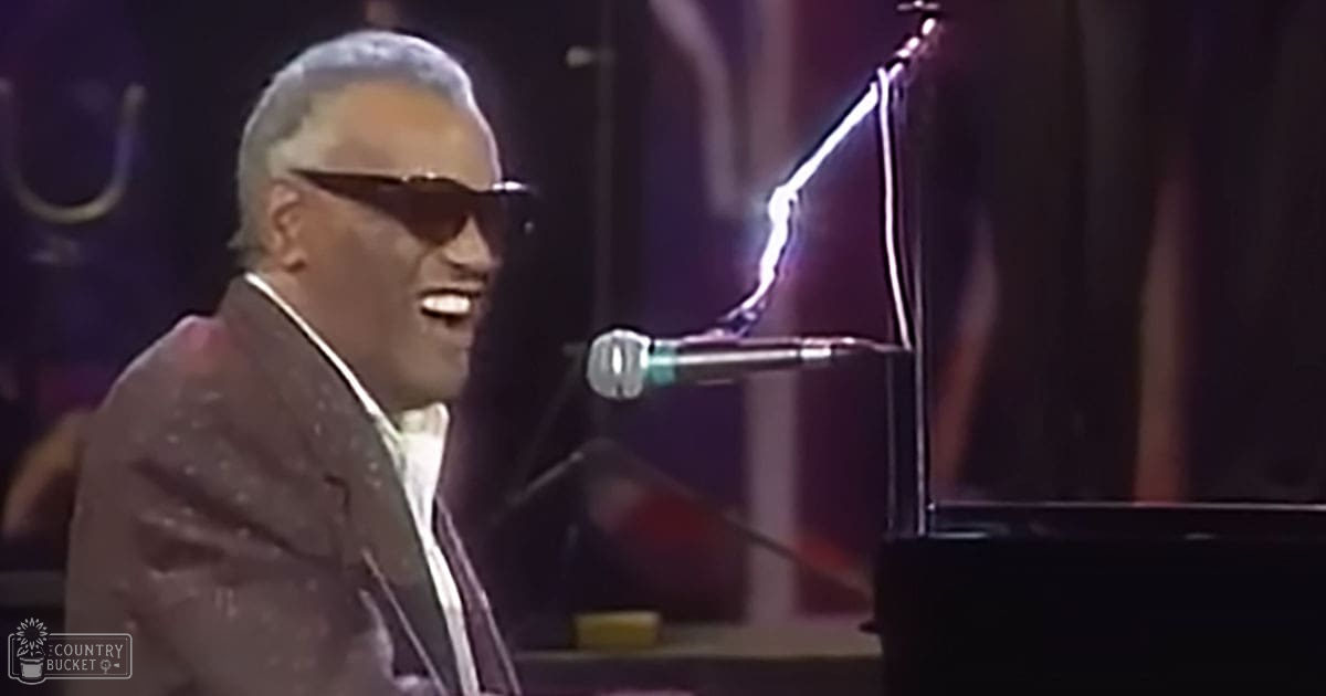 An older adult male singer wearing a dark brown suit jacket with a subtle pattern and white shirt, performing at an indoor concert venue.