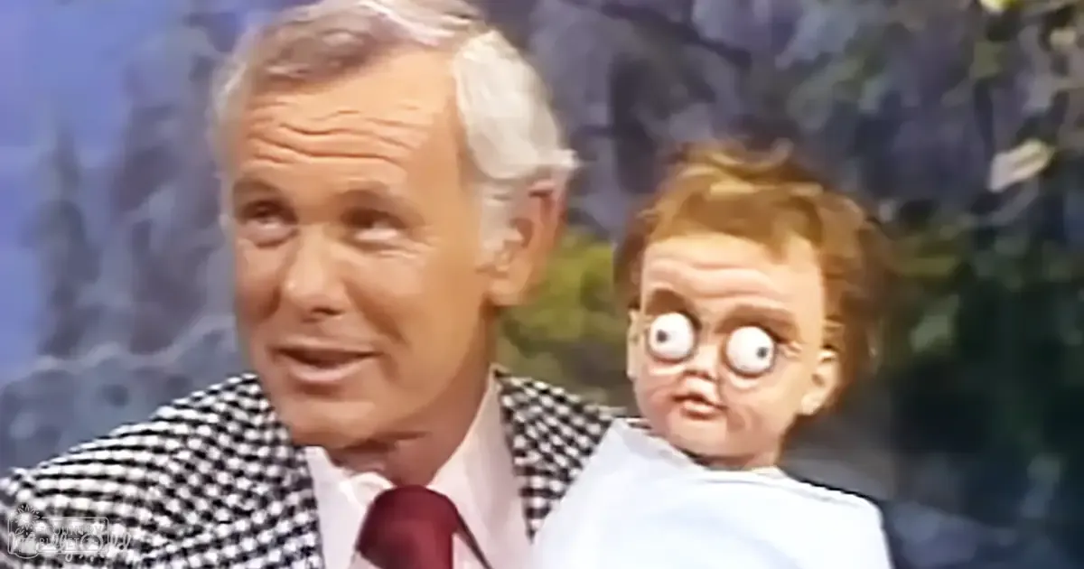 Middle-aged adult male holding an infant wearing a checkered suit jacket with white shirt and red tie, likely from the 1950s or 1960s in a television studio setting.