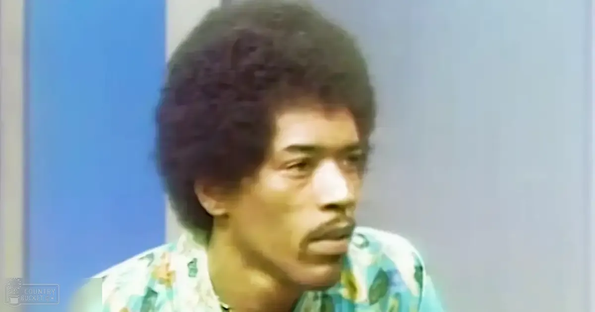 A young adult male wearing a loose, collared Hawaiian-style shirt with green floral patterns stands against a simple blue wall indoors.