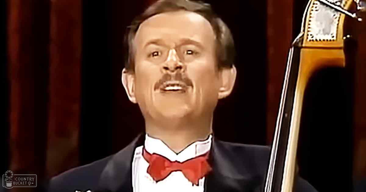 Adult male singer in a mid-20th century black suit with a white shirt and red bow tie, holding a double bass on an indoor stage with dark curtains as backdrop.