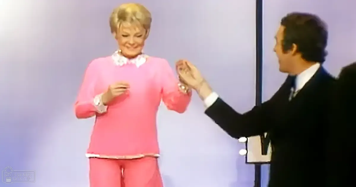 An older adult female in a pink sweater with a small heart design, paired with high-waisted pants at what appears to be an indoor event. An adult male wearing a dark suit holds a small object.