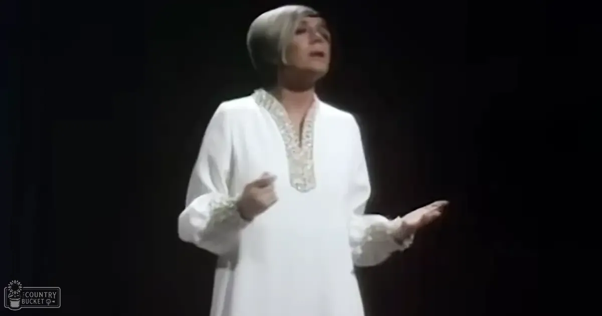 A middle-aged woman performs on a dark stage, wearing a loose-fitting long-sleeved white garment with intricate embroidery around the neckline and cuffs.