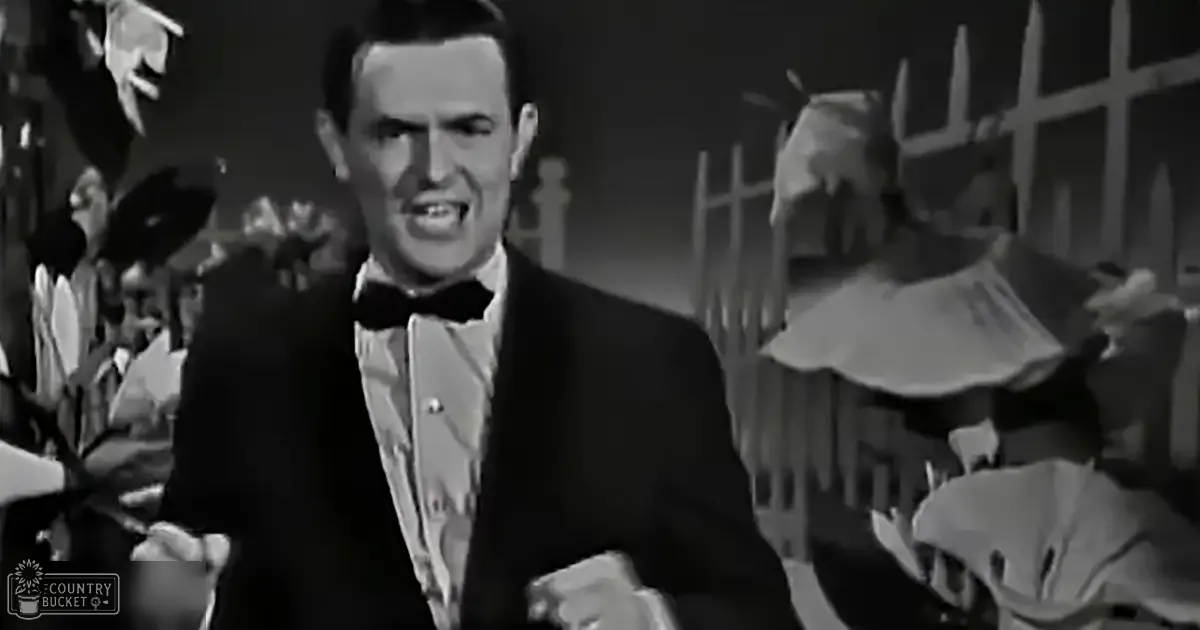 A man wearing a formal tuxedo with a bow tie stands on stage, surrounded by decorative elements like flowers and a fence, suggesting an outdoor or semi-outdoor venue during the day.