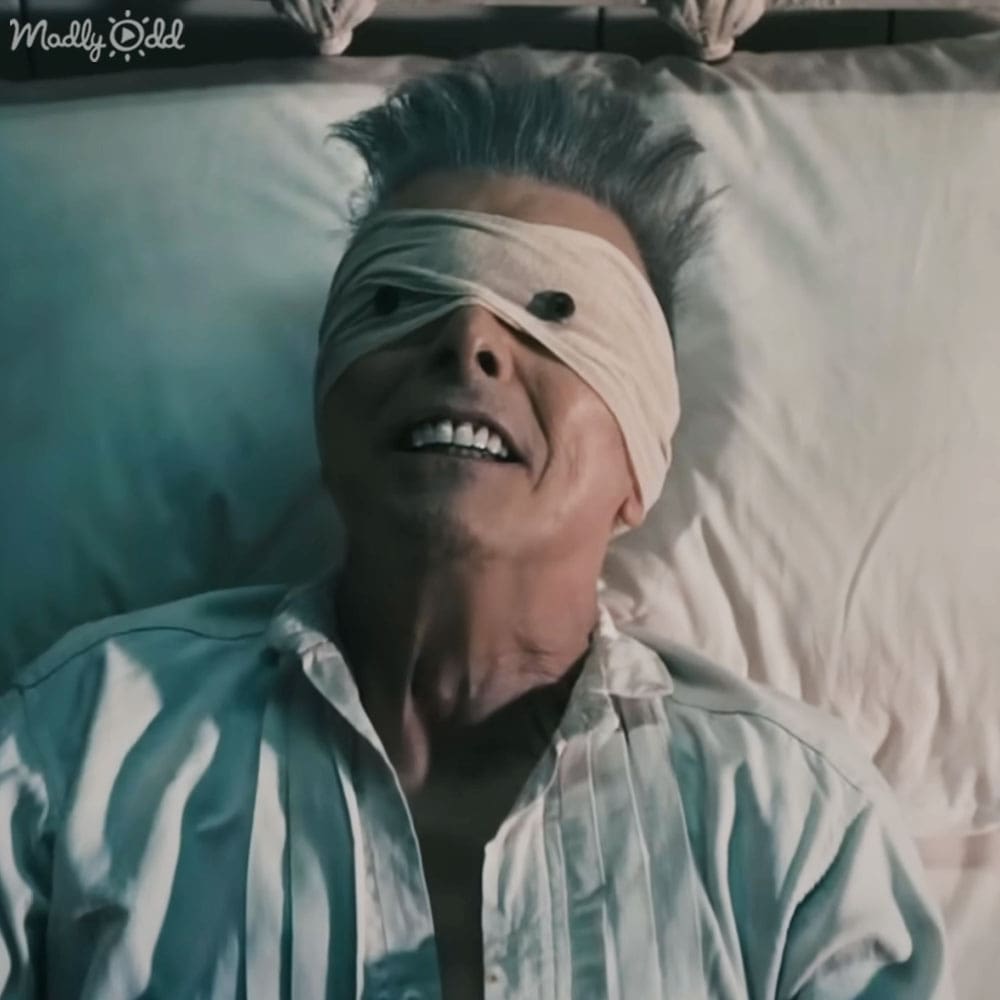 A middle-aged man lies in bed, wearing a relaxed-fit striped button-up shirt with white and teal horizontal stripes, likely in a modern bedroom setting under natural light.