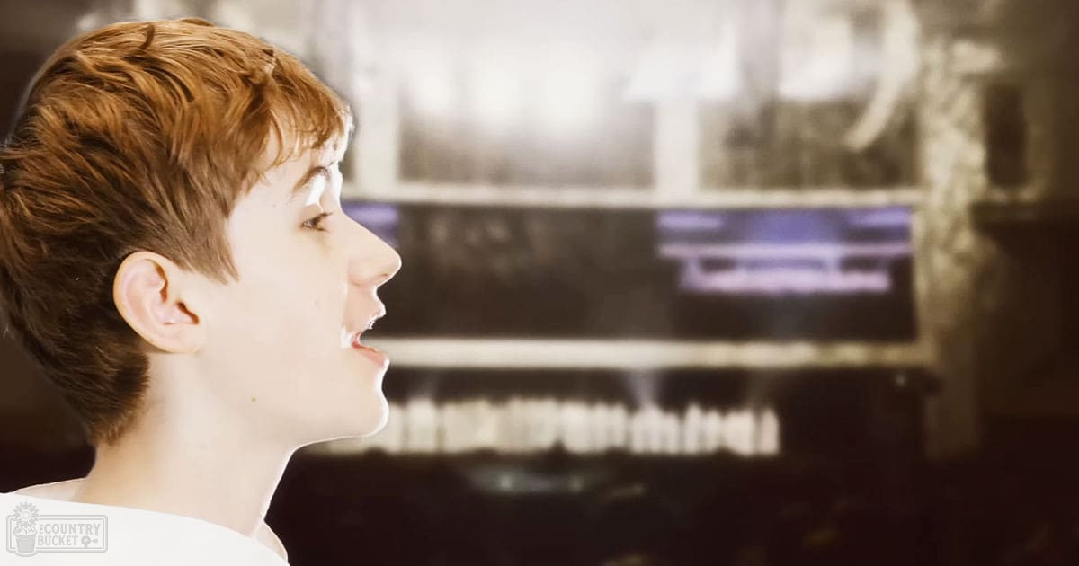 A young child with short brown hair wearing a loose-fitting white shirt or tunic stands indoors near a grand piano under warm artificial lighting, giving off a vintage sepia tone.