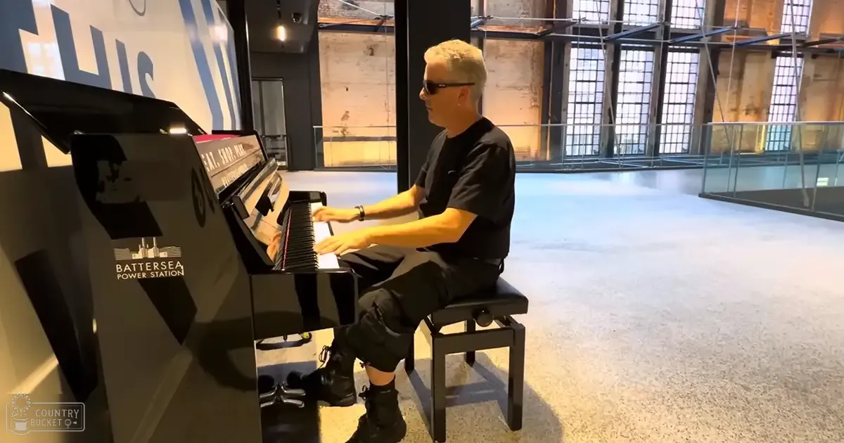 Adult male playing piano at Battersea Power Station with black T-shirt, shorts, boots, and sunglasses in an indoor space with high ceilings and industrial-style windows.