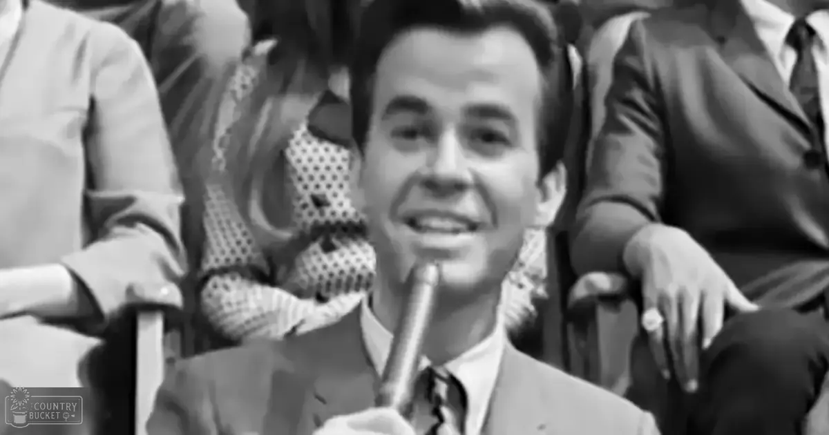 An indoor setting with a young adult male in formal attire from the mid-20th century seated among other individuals at what appears to be an event or gathering, possibly during spring or summer.