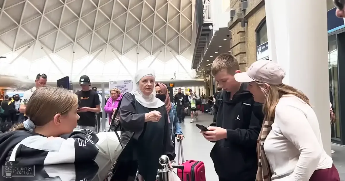 A nun interacts with a group of people in an indoor public space, possibly a shopping mall or train station. A child wears a black and white hoodie, while others wear casual clothing like hoodies and jackets.