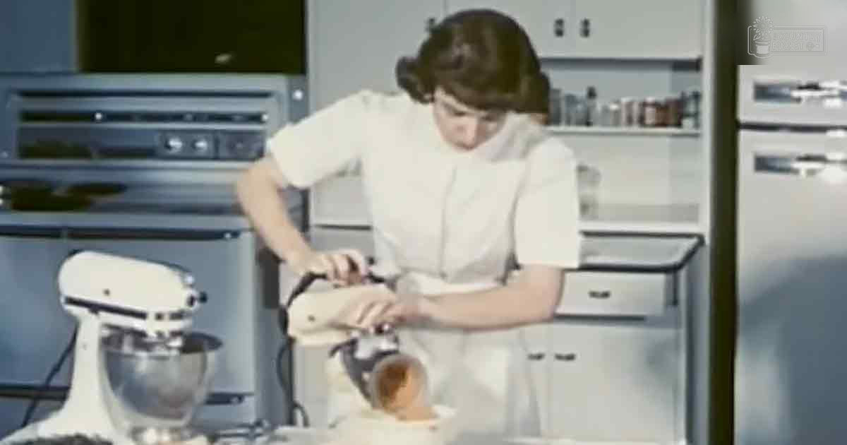 A young adult female wearing a white apron in a mid-20th-century style kitchen setting with beige cabinets, silver appliances, and light daytime lighting.