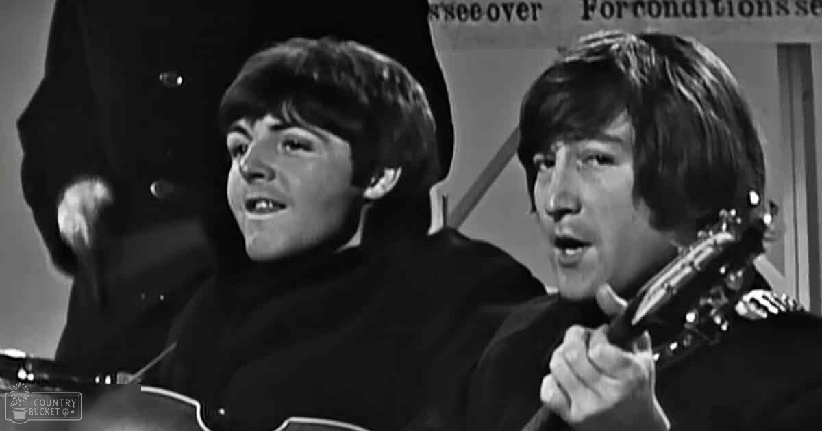 Two young adult males performing on stage, wearing dark turtleneck sweaters from the mid-20th century, holding guitars in a live performance setting with no visible natural light or animals.