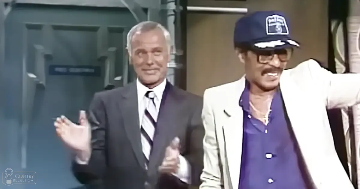 Two middle-aged men in a studio setting with comedic attire; one wears a dark suit with a striped tie, the other a white blazer over a purple shirt and a dark blue cap.