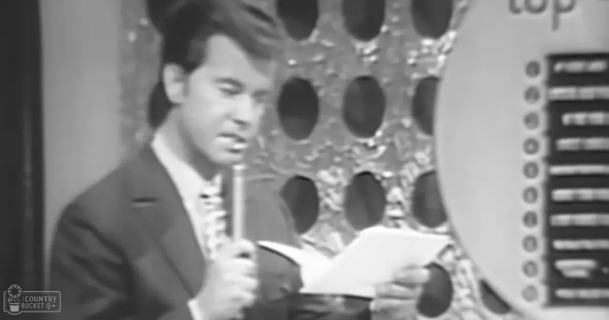 A young adult male in a mid-20th-century style dark suit jacket with a patterned tie stands indoors, possibly in a studio setting, holding a microphone and a piece of paper.