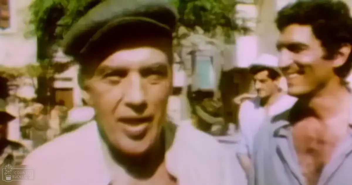 An older adult male wearing a white shirt and dark cap smiles outdoors in what appears to be a town square with trees and buildings, late afternoon lighting.