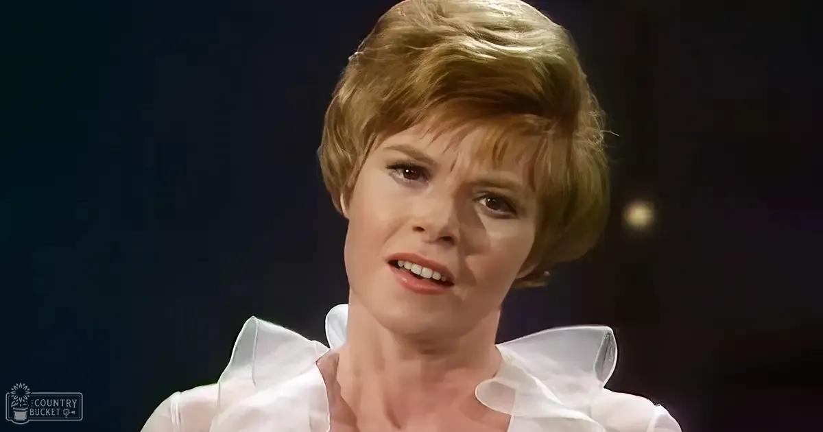 A middle-aged woman wearing a white blouse with puffed shoulders and ruffled neckline, possibly from the mid-20th century, stands against a dark background in what appears to be a formal or professional setting.