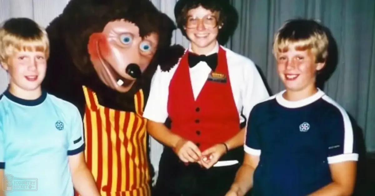 A mascot character resembling an animal, flanked by two children with an adult behind them in a school event setting; light blue and dark trim shirts on the children, striped yellow and orange shirt on the mascot, white shirt under red vest on the adult; plain curtain background.