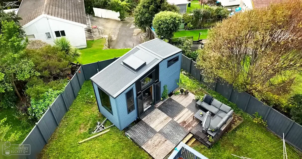 A modern tiny house with large windows and solar panel sits in a residential backyard surrounded by greenery, suggesting spring or summer day.